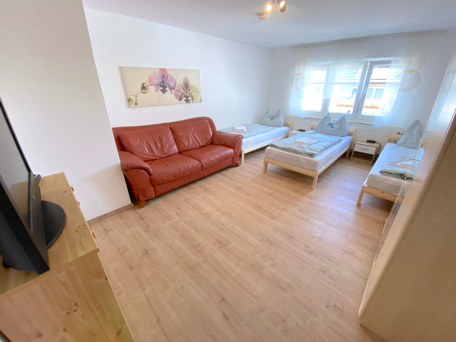 Apartment IV in Kronwieden – Großer Raum mit drei Betten, rotem Ledersofa und Fenstern